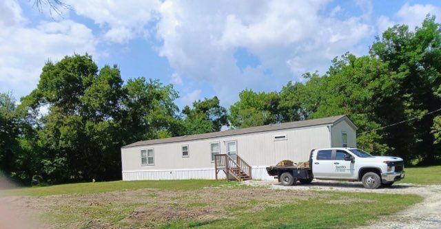 White mobile home in a grassy lot with a pickup truck parked outside, surrounded by trees and blue sky.