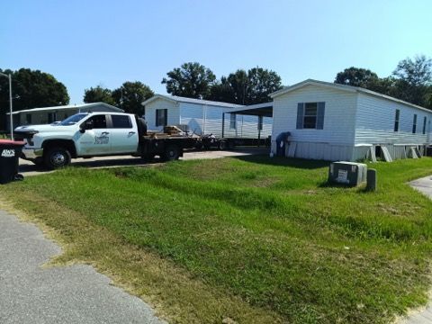 White pickup truck and flatbed trailer parked beside a small white house on a sunny lawn.