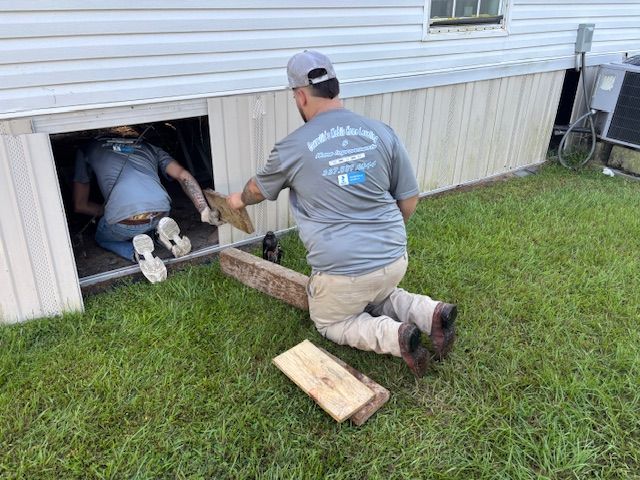 Two men working under a house: one crawls inside, the other kneels handing him wood on grass.
