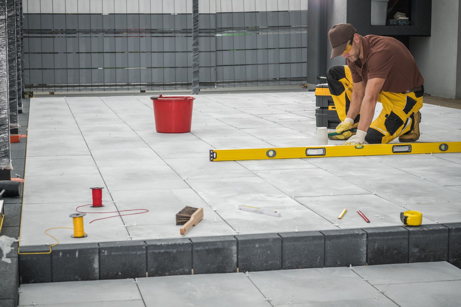 Man in work clothes laying paving stones with level, red bucket nearby.