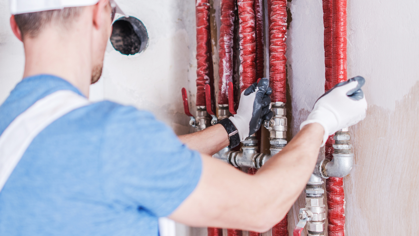 Plumber in blue shirt and gloves inspects red insulated pipes with valves in a room.