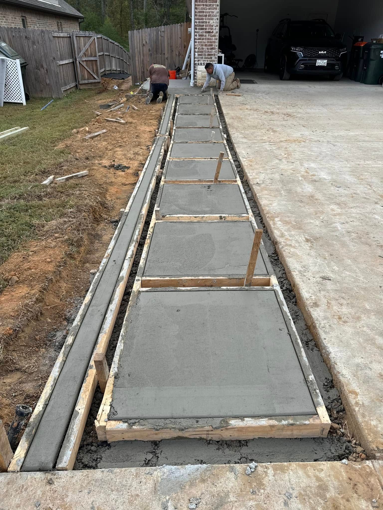 A view looking down a new concrete walkway under construction alongside a paved driveway and a wooden fence.