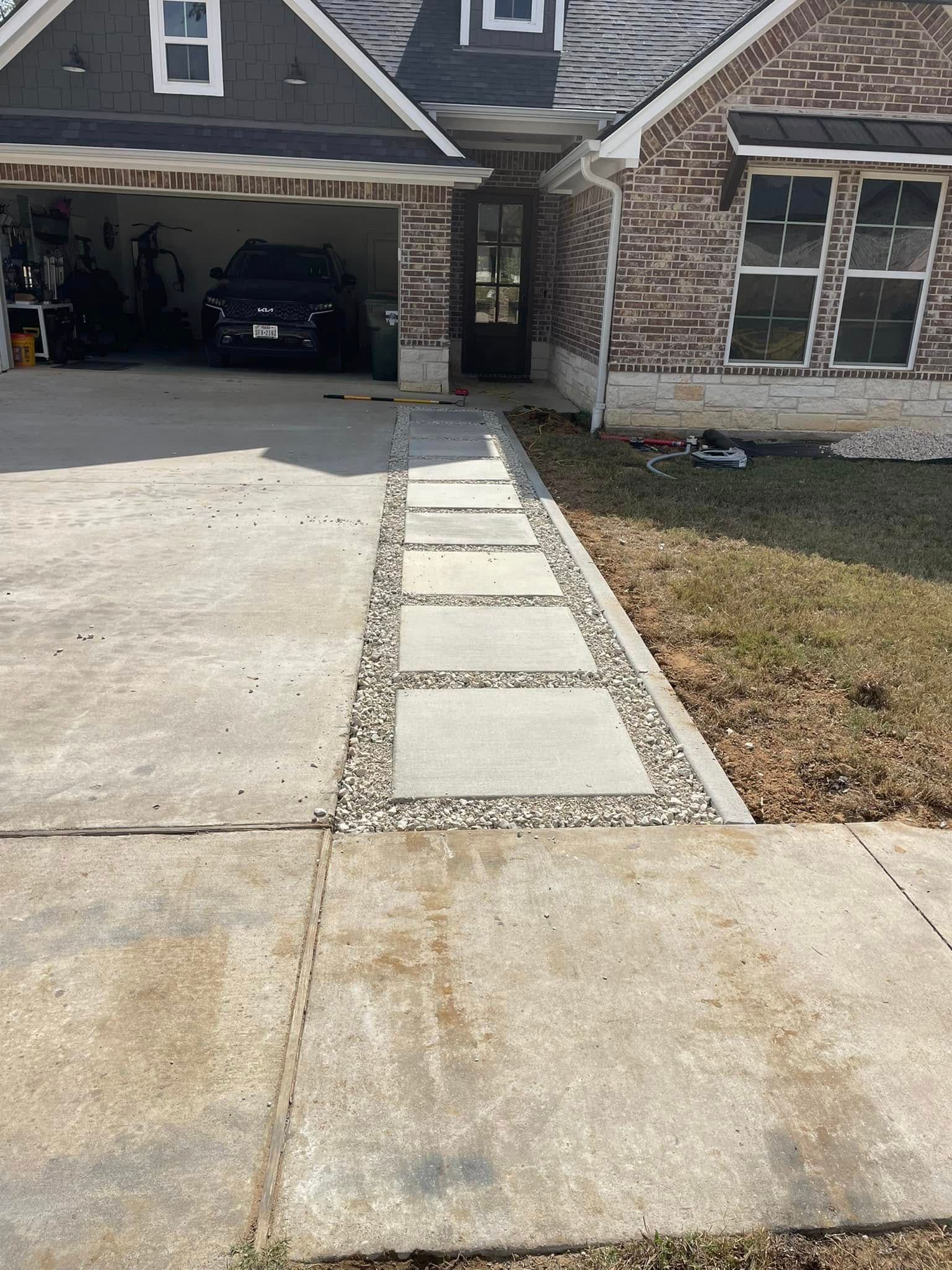A concrete walkway with square pavers and gravel borders leads to a suburban brick house with a garage.