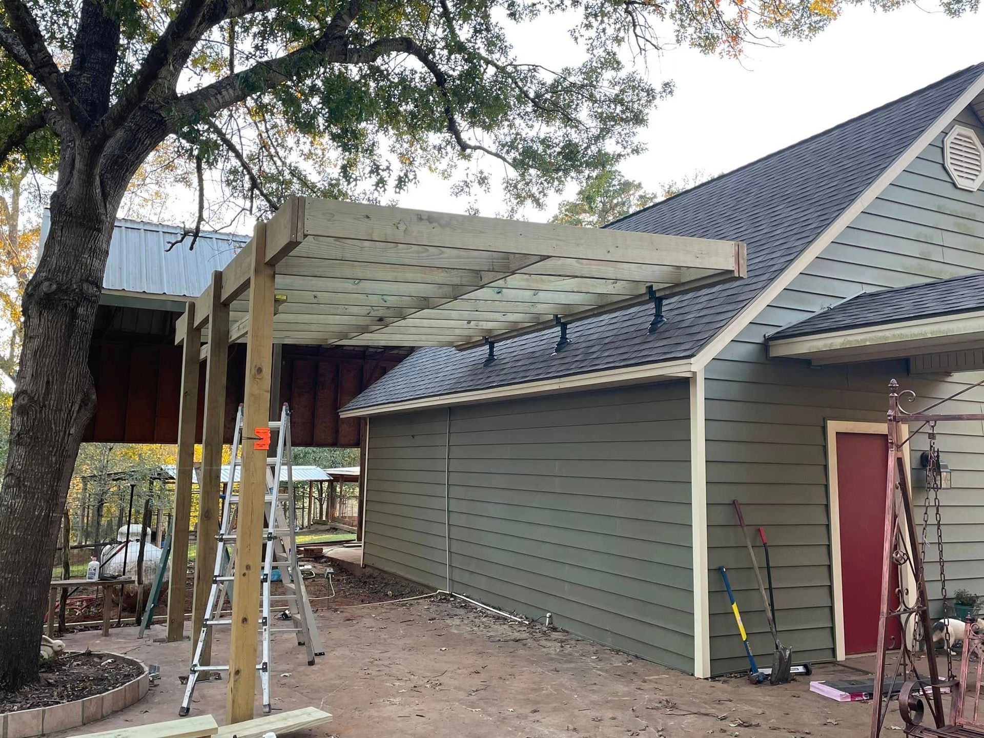 A wooden pergola structure under construction attached to the side of a grey-sided house with a dark metal roof.