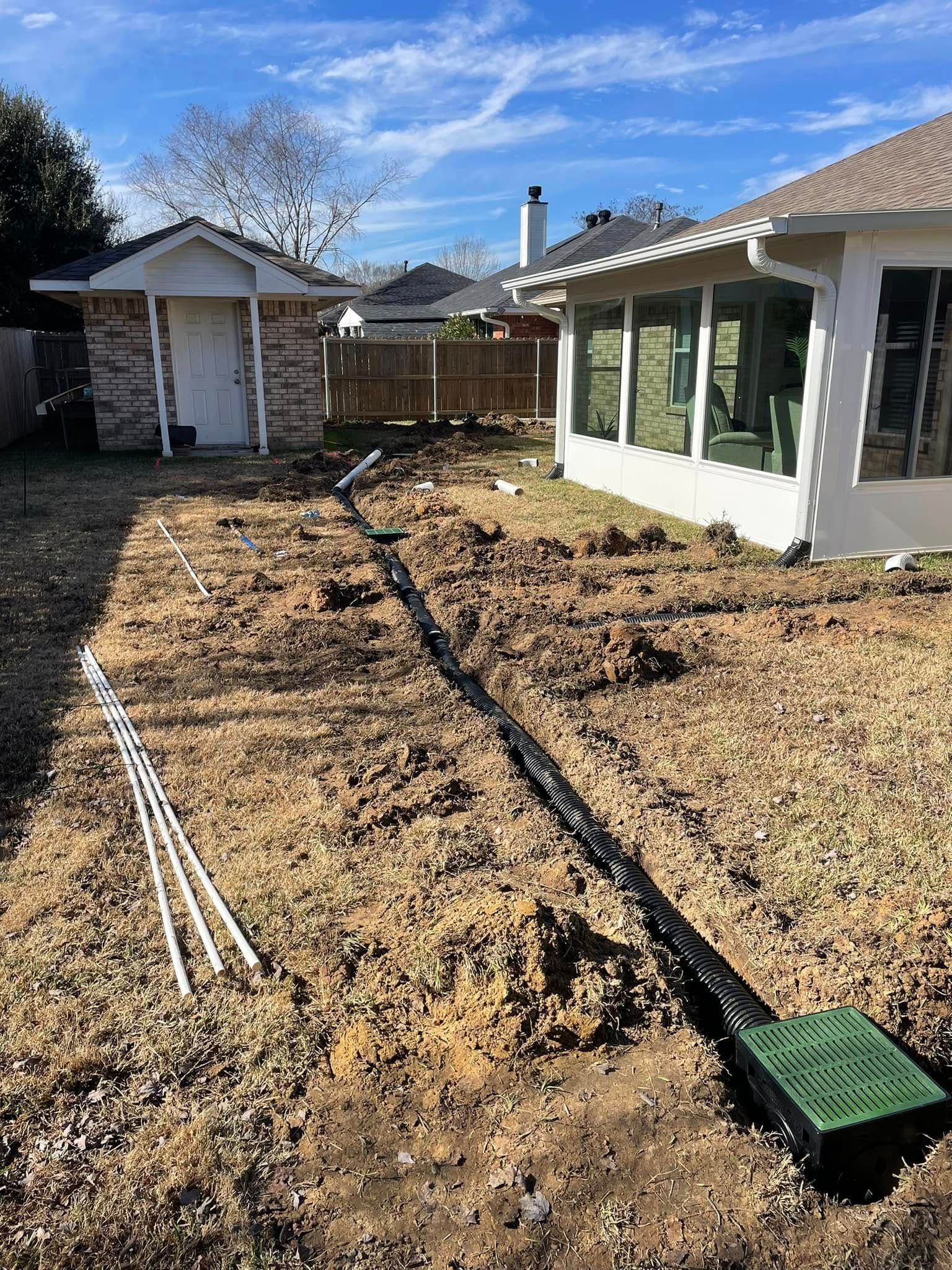 A black corrugated drainage pipe connects a green catch basin in a yard under renovation, near a house and a shed.