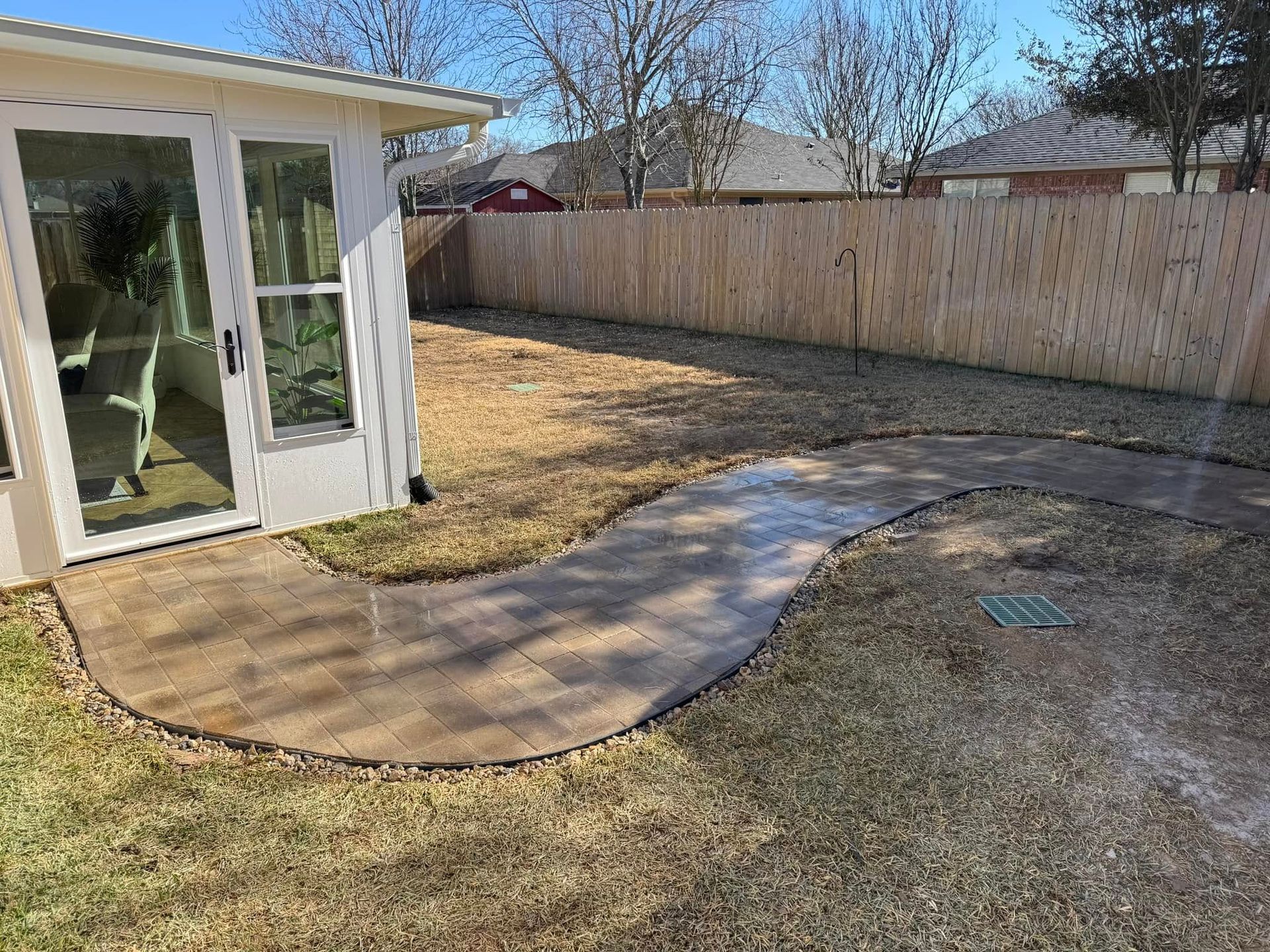 A curved paver walkway leads from the side of a white structure into a backyard with a wooden fence.