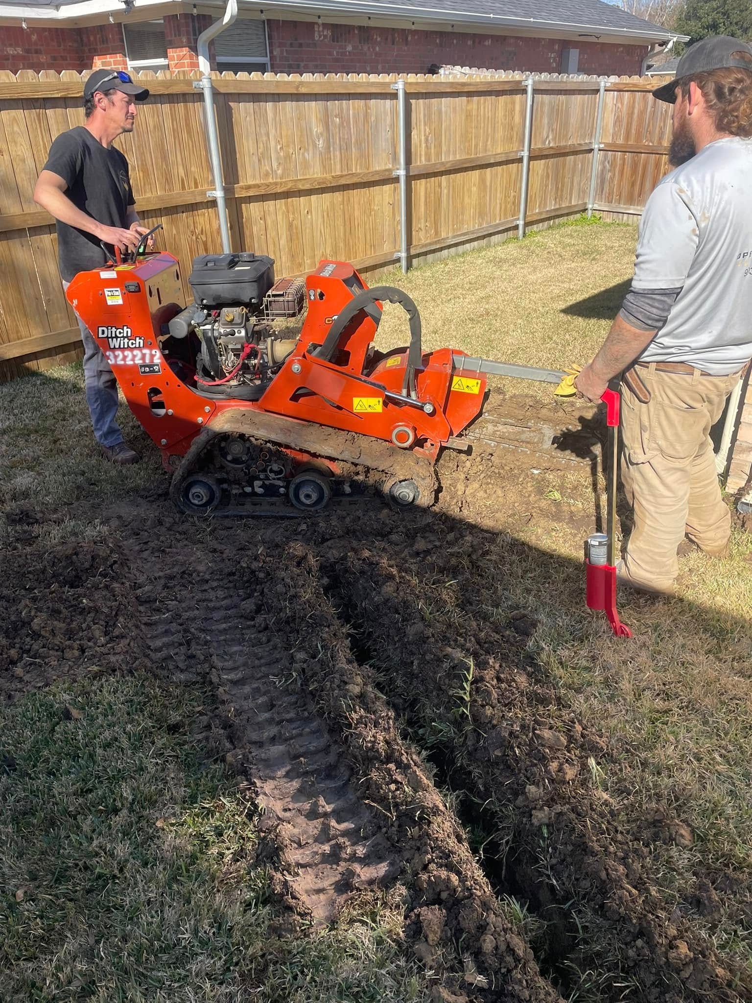 Two people in a grassy yard operate an orange trencher machine to dig a long, narrow trench through the soil.