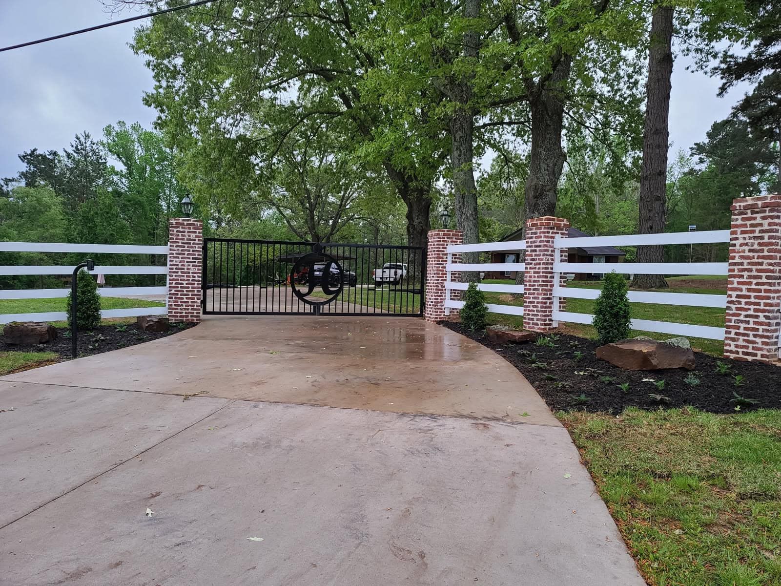 A black metal gate with a circular monogram sits between brick pillars and white split-rail fencing on a driveway.