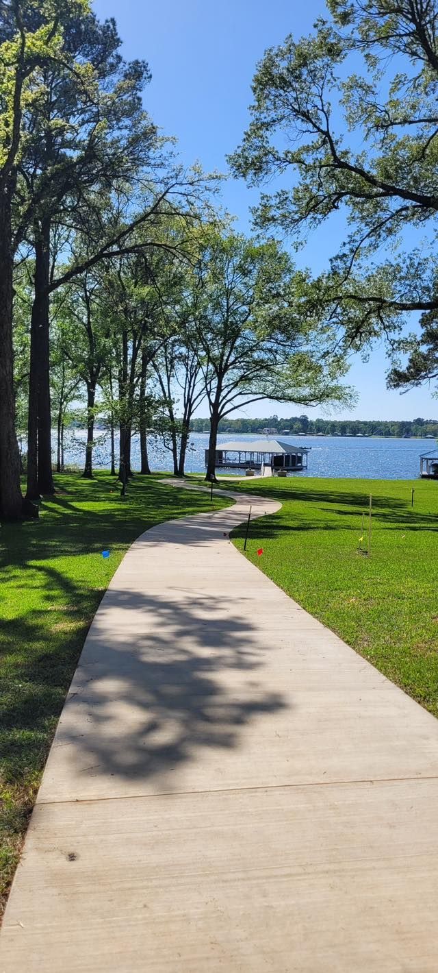 A concrete path winds through a grassy, tree-lined park toward a lake with a boat dock under a clear blue sky.