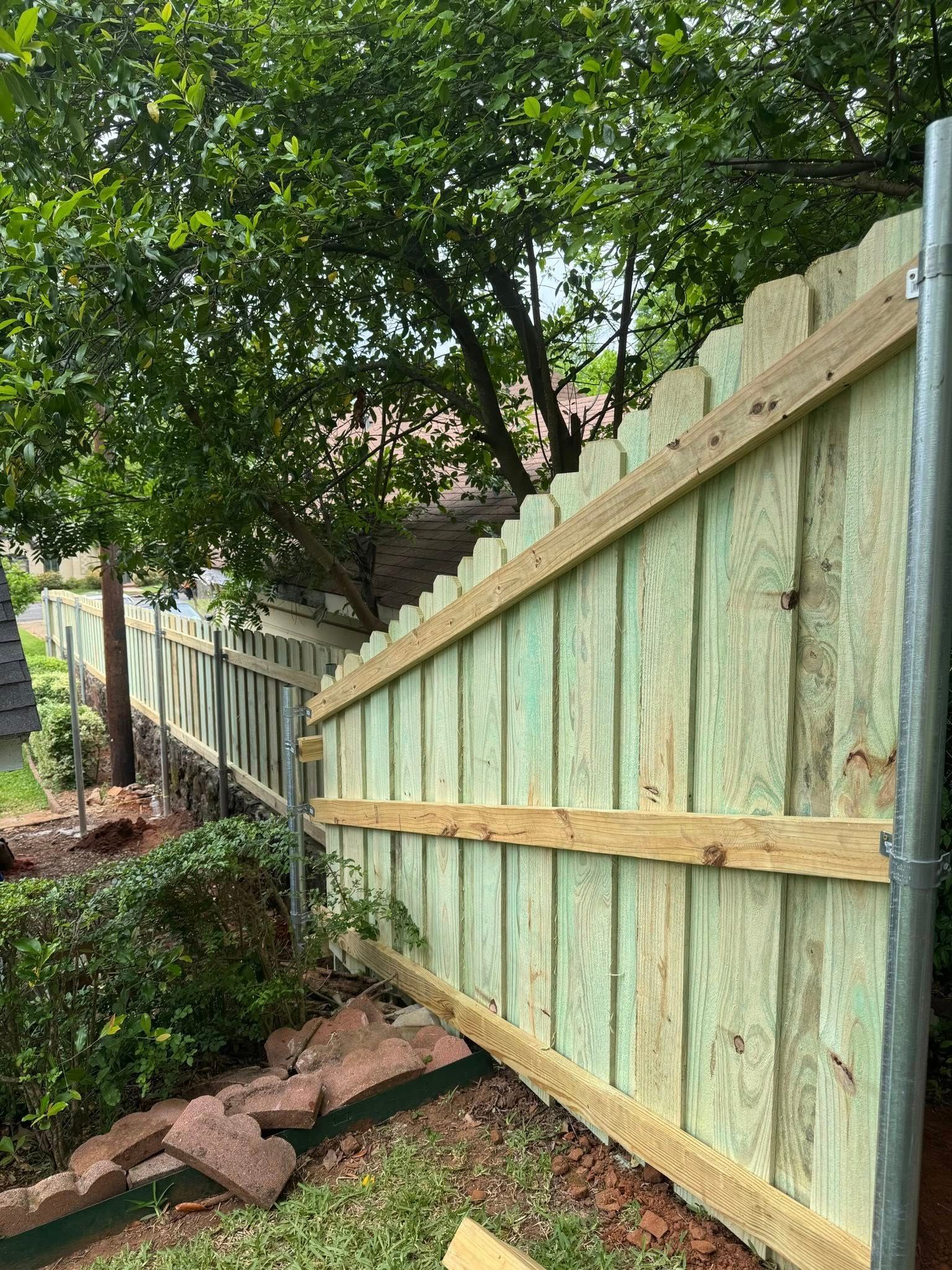 A new, light-green wooden fence section installed at an angle with metal posts in a yard with a tree and landscaping.