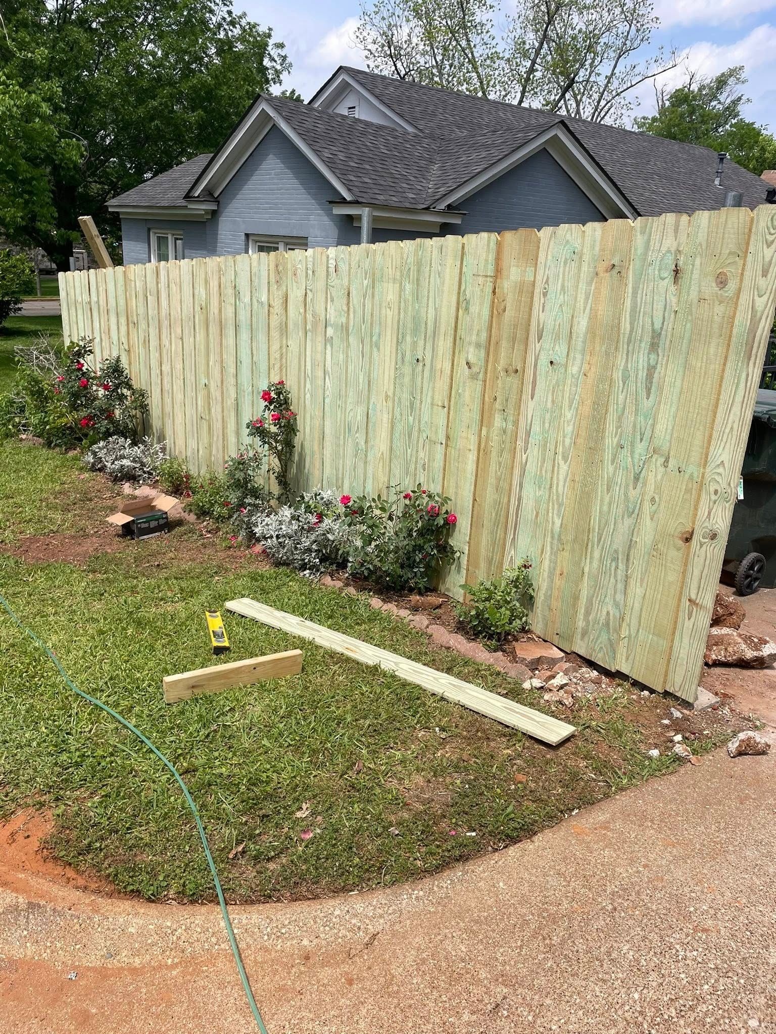 A new, light-green wooden fence stands in a yard in front of a blue house, with landscaping and tools on the grass.