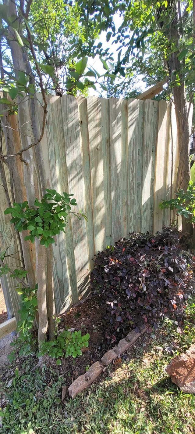 A vertical wooden fence framed by trees, with a small bush and a brick-lined garden bed in the foreground.