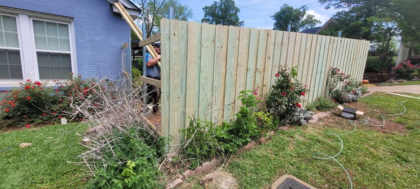 A worker installing a light-colored wooden privacy fence next to a blue house with blooming flower bushes in the yard.