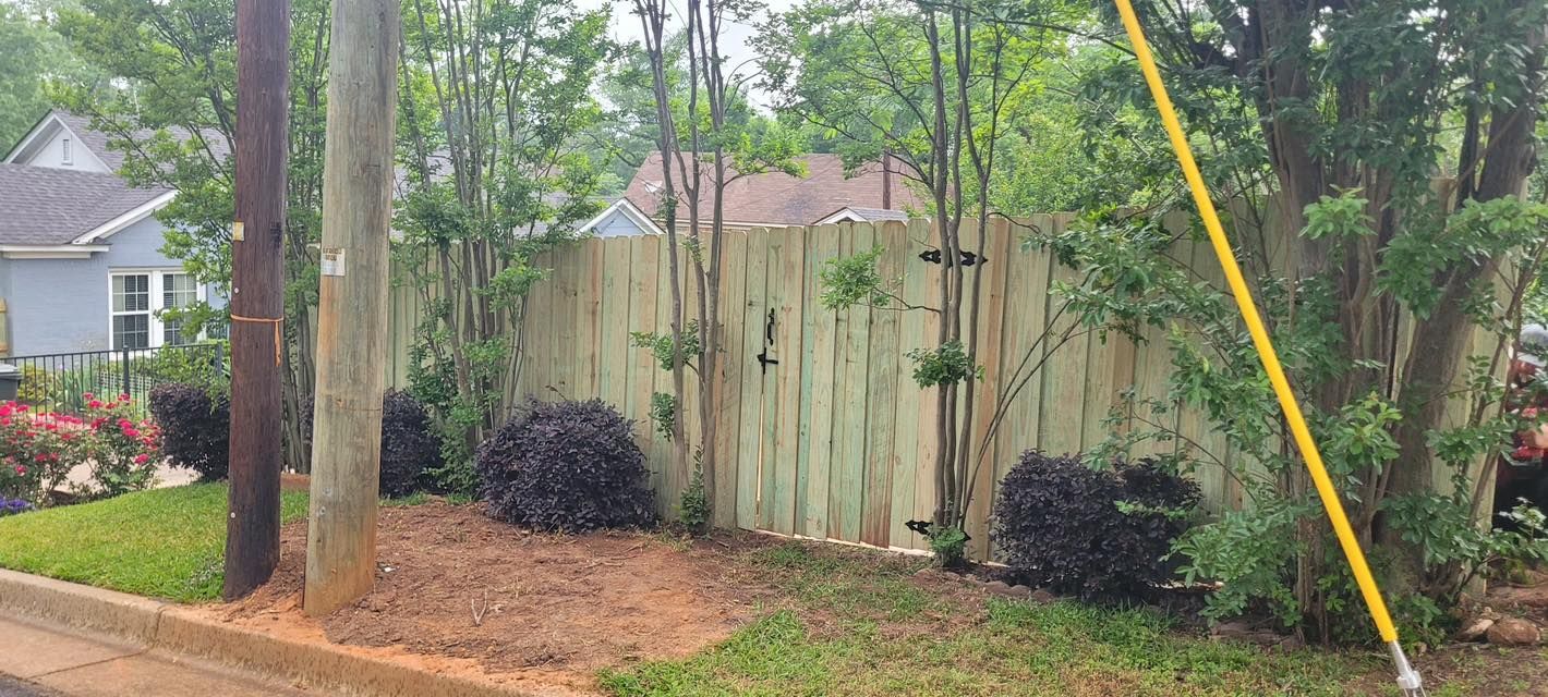 A wooden fence stands behind manicured shrubs and trees in a residential neighborhood.