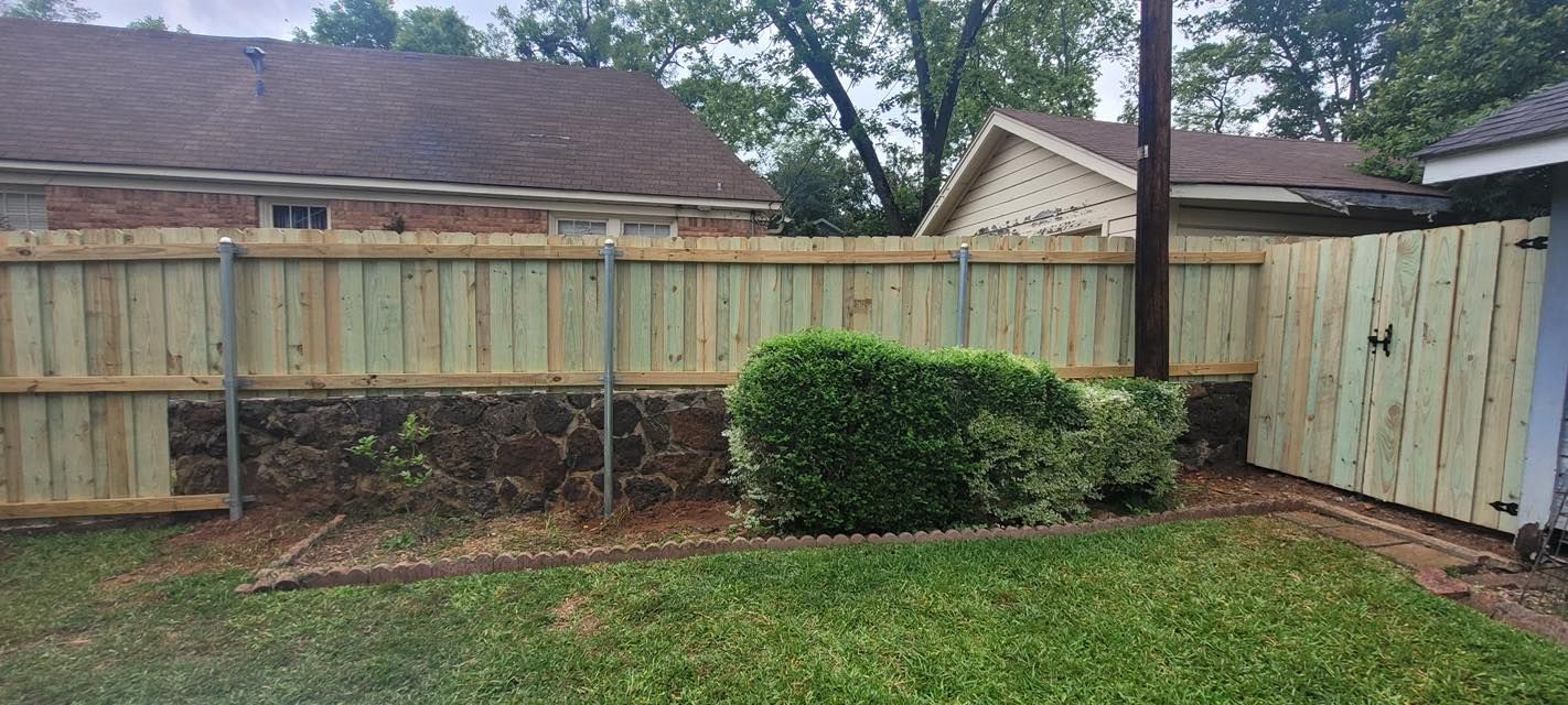 A new wooden privacy fence featuring a section of stone base, with a green bush and a manicured lawn in the foreground.