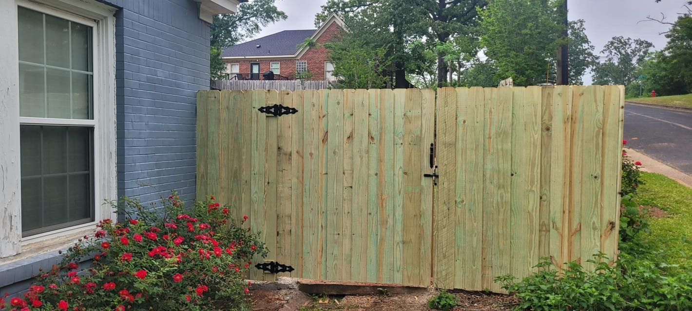 A wooden gate attached to a gray brick house exterior next to a bush with red flowers.
