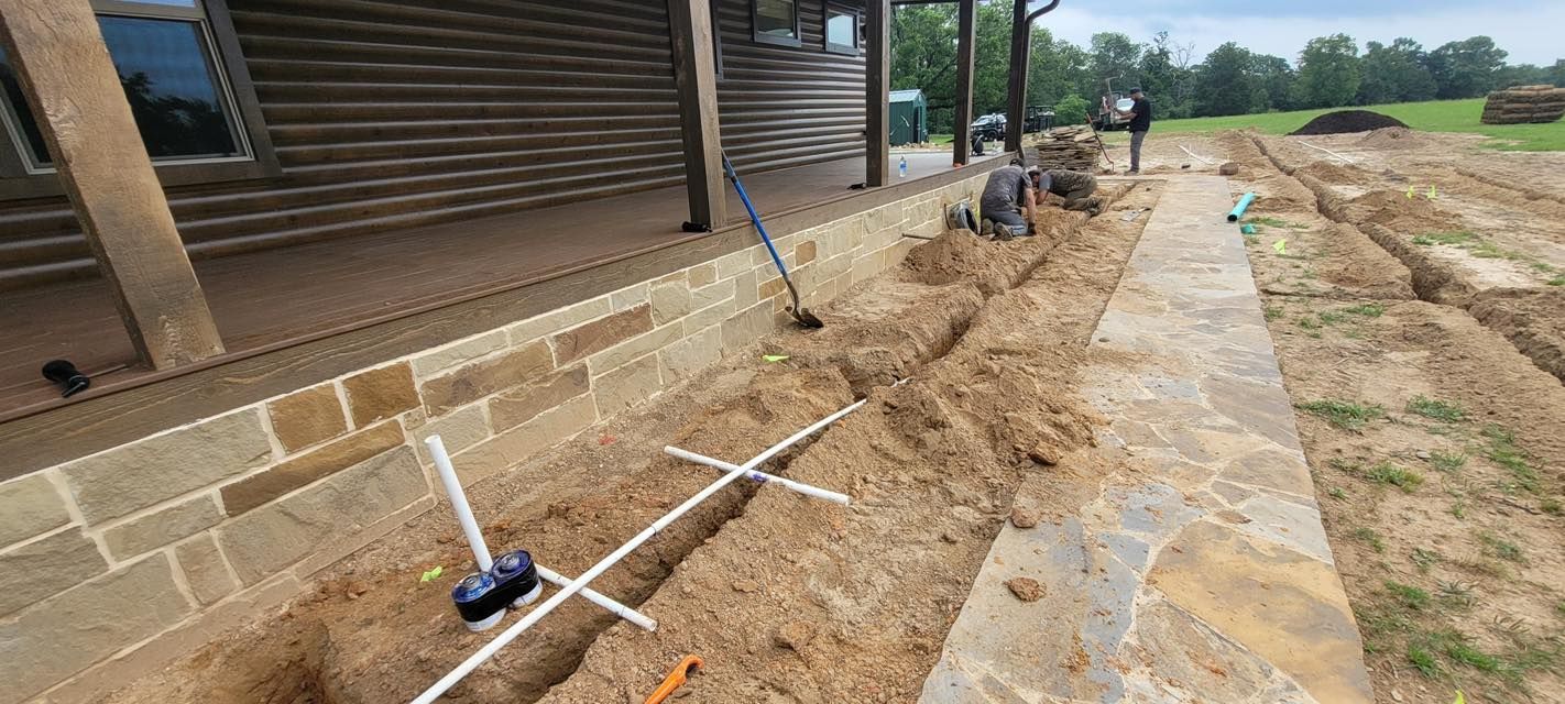 A trench with white PVC pipes being installed along the stone foundation of a log cabin with a stone patio alongside.