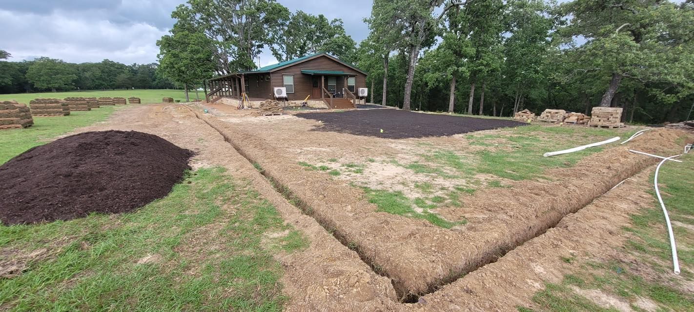 A cabin sits in a yard with a large pile of dark mulch, trenches, and prepared garden beds under construction.