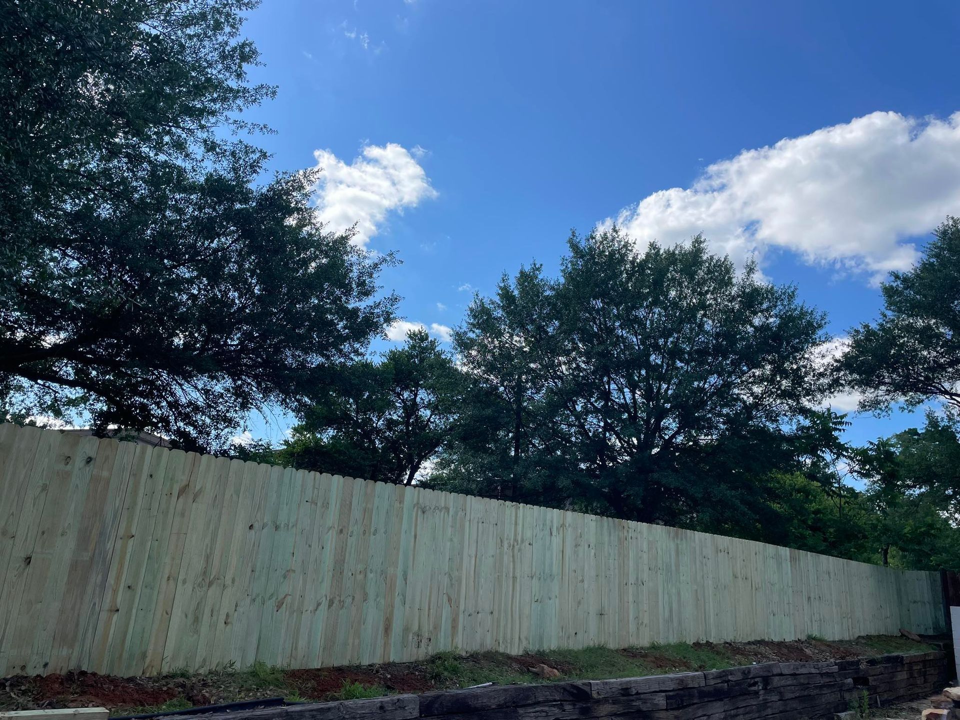 A new wooden fence stands in front of lush green trees under a bright blue sky with scattered white clouds.