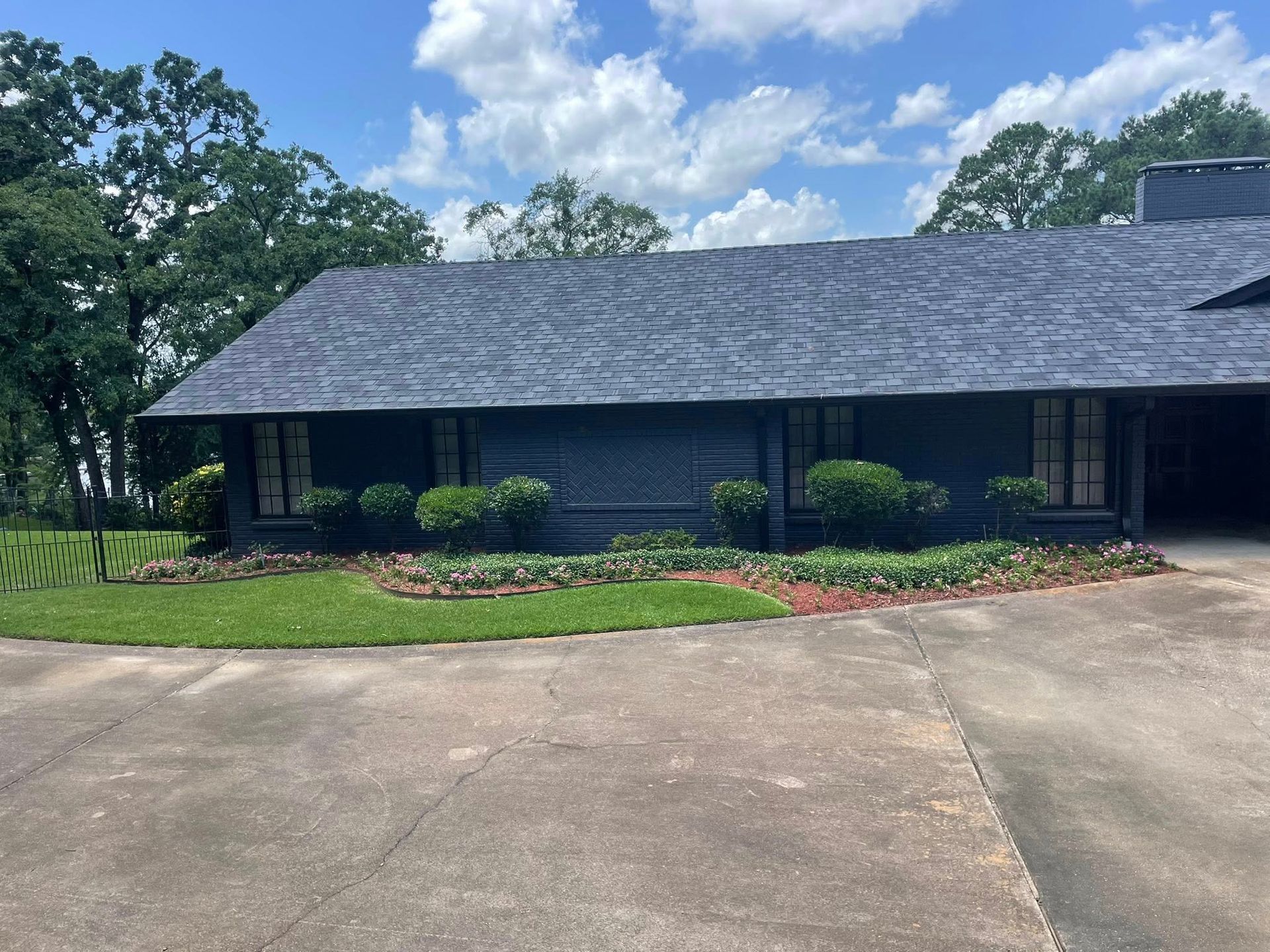 A one-story dark gray house with a large roof sits behind a concrete driveway and a manicured green lawn under a blue sky.