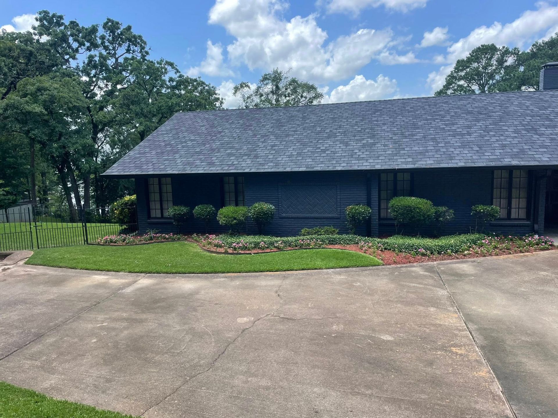 A dark-colored, single-story house with a shingled roof, surrounded by green shrubs and trees under a blue sky.