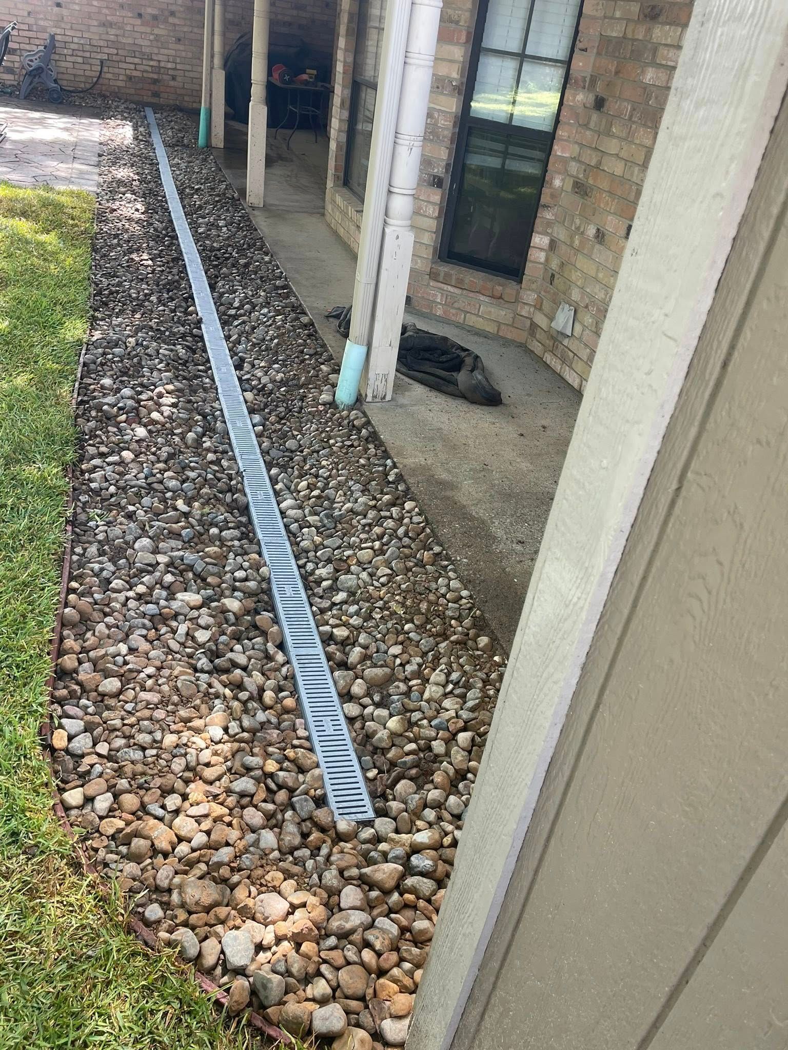 A long, narrow gray metal drainage grate installed in a bed of decorative river rocks along a brick house exterior.