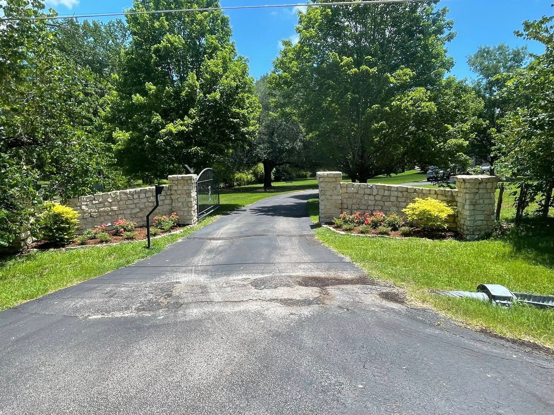 A stone-pillared driveway entrance lined with green trees and small flowering bushes under a bright blue sky.