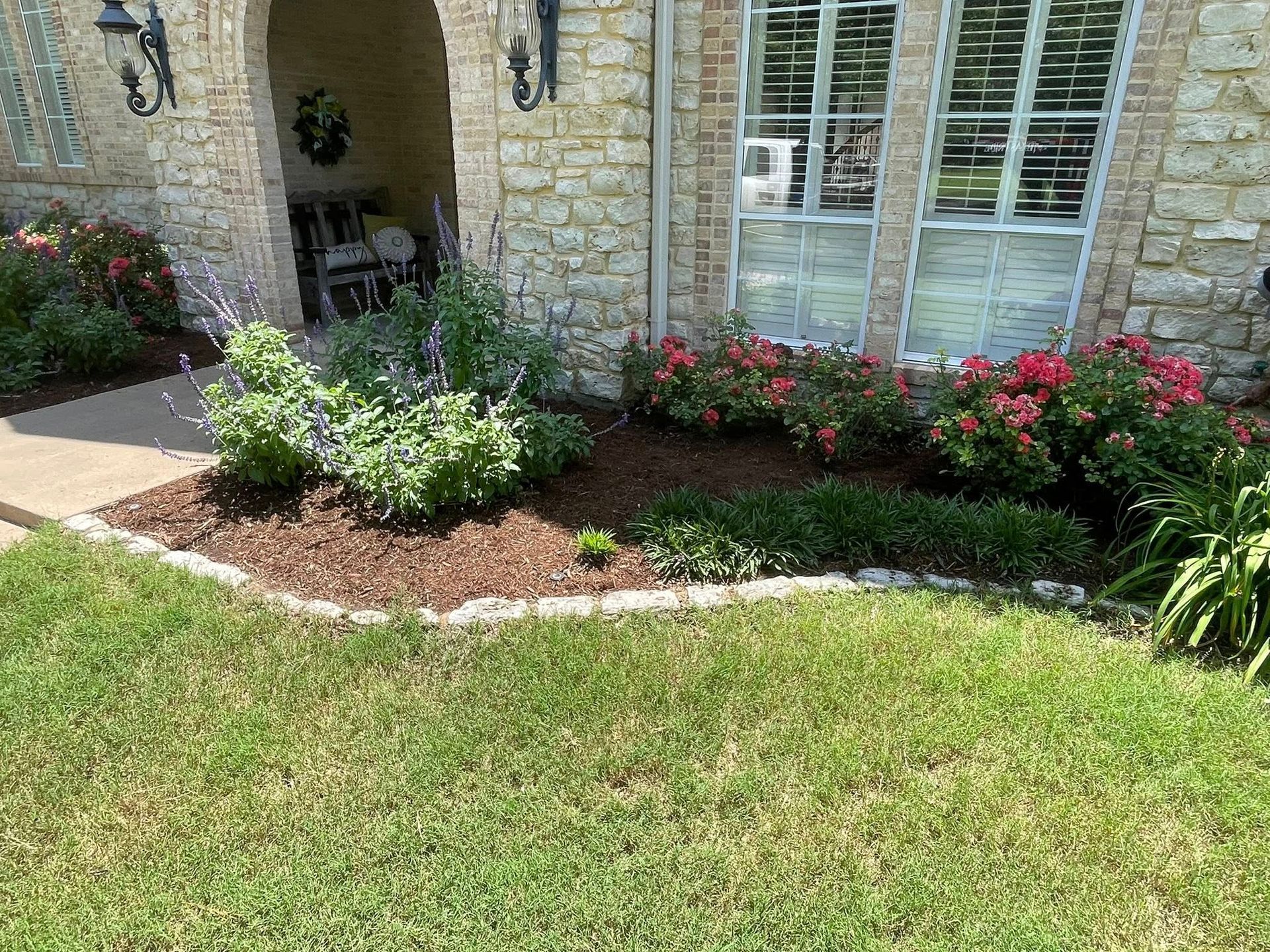 A stone house exterior features a shaded entryway, a window with white shutters, and a flowerbed filled with blooming roses.