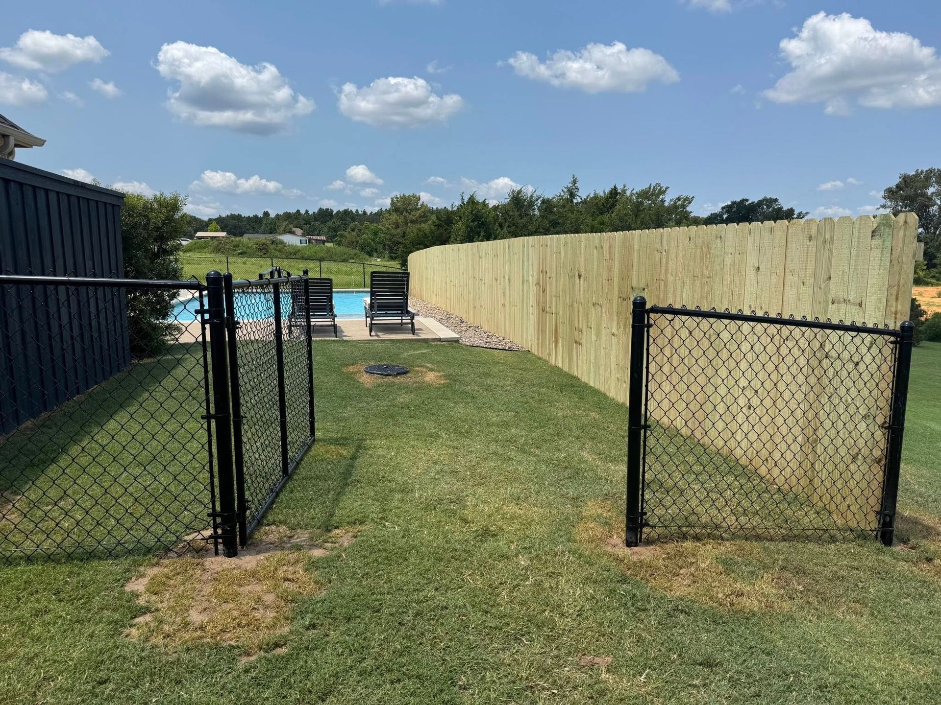A grassy backyard features a short black chain-link gate opening towards a swimming pool, bordered by a tall wooden fence.