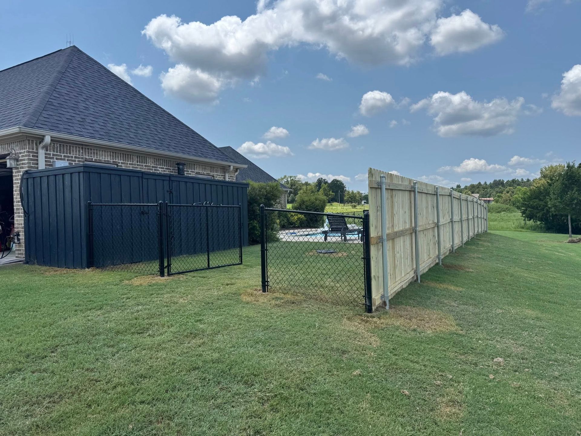 A dark blue storage container sits next to a wooden fence and a black chain-link gate in a grassy yard under a blue sky.