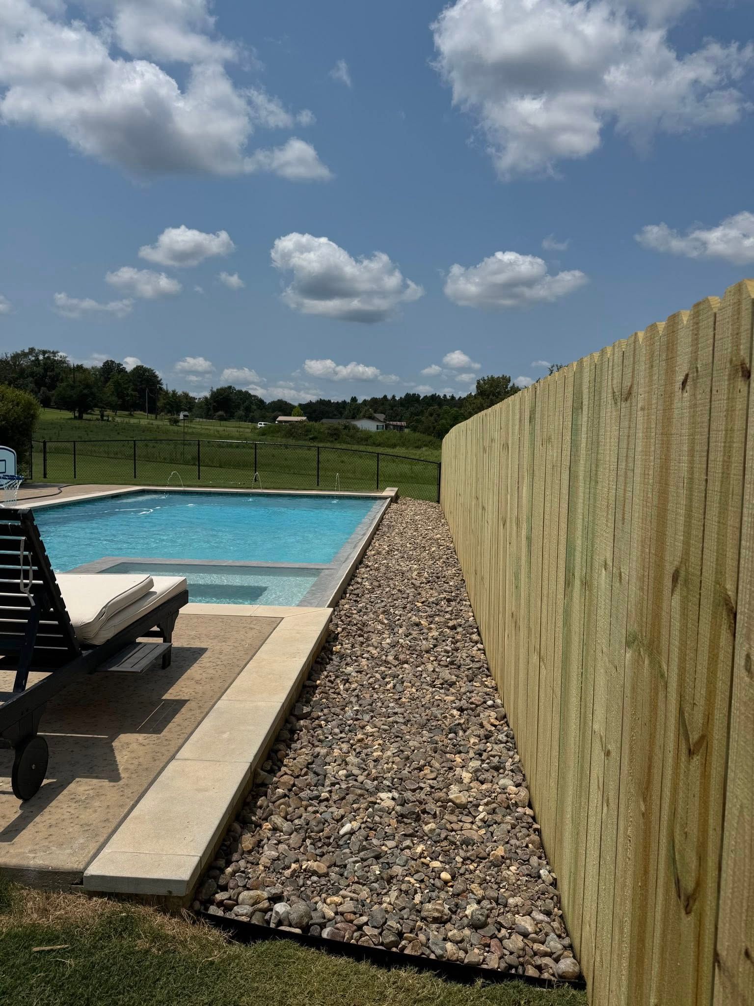 A blue swimming pool sits next to a gravel strip and a tall wooden privacy fence under a sunny, partly cloudy sky.