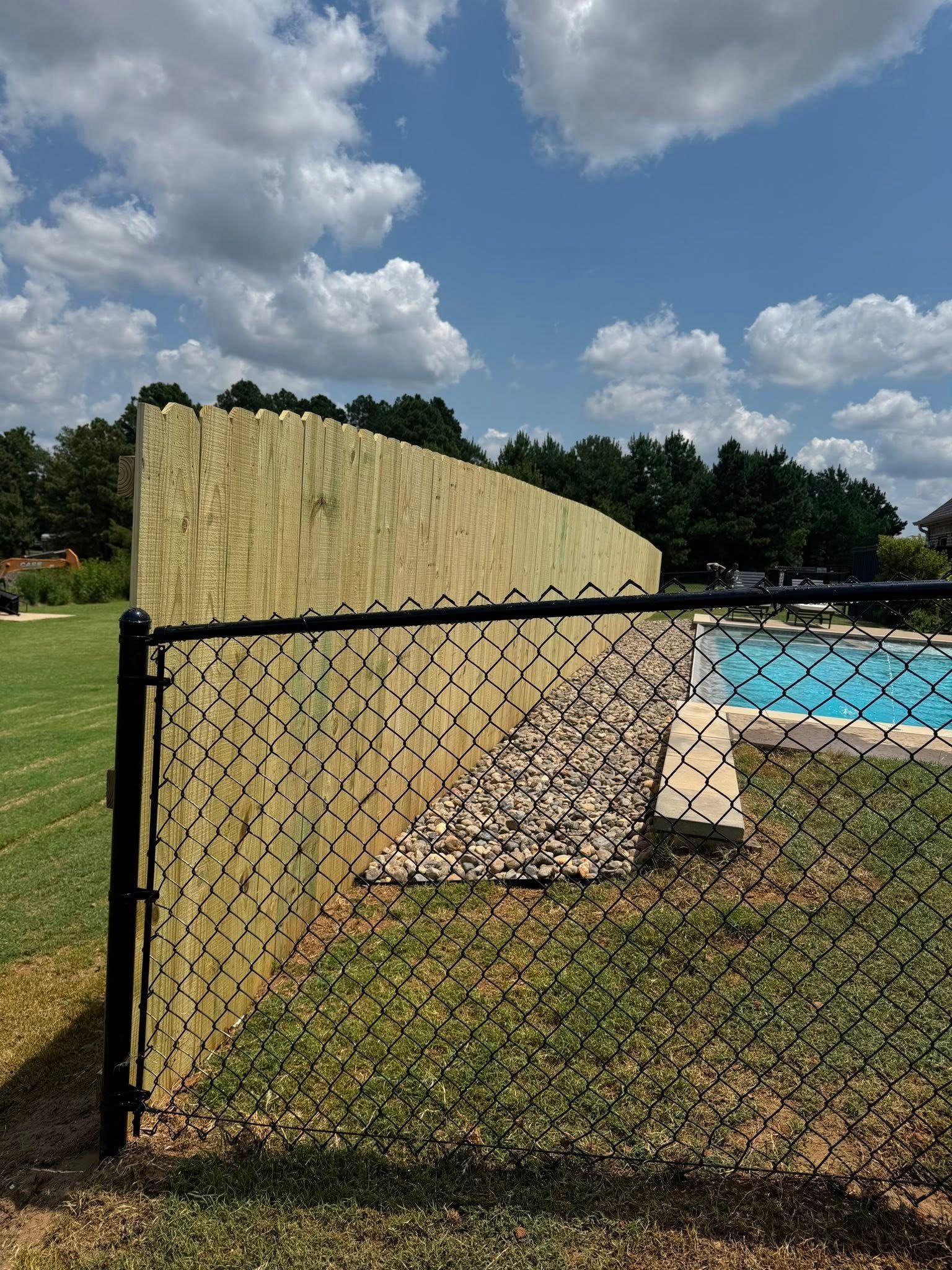 A wooden privacy fence stands behind a black chain-link fence, with a rocky landscaping border and a swimming pool nearby.