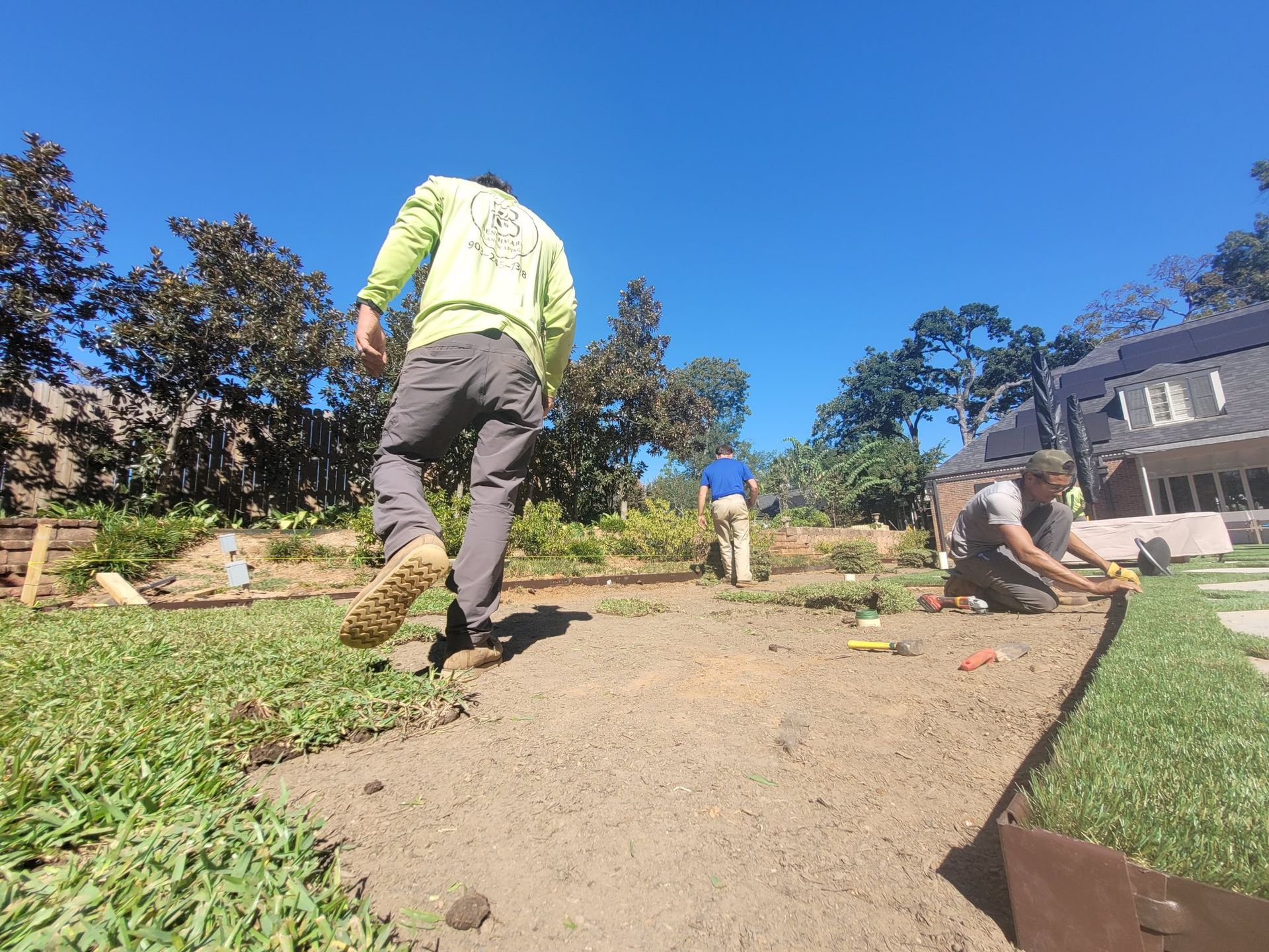 Three landscapers work on a lawn project, installing metal edging and preparing soil in a sunny outdoor yard.