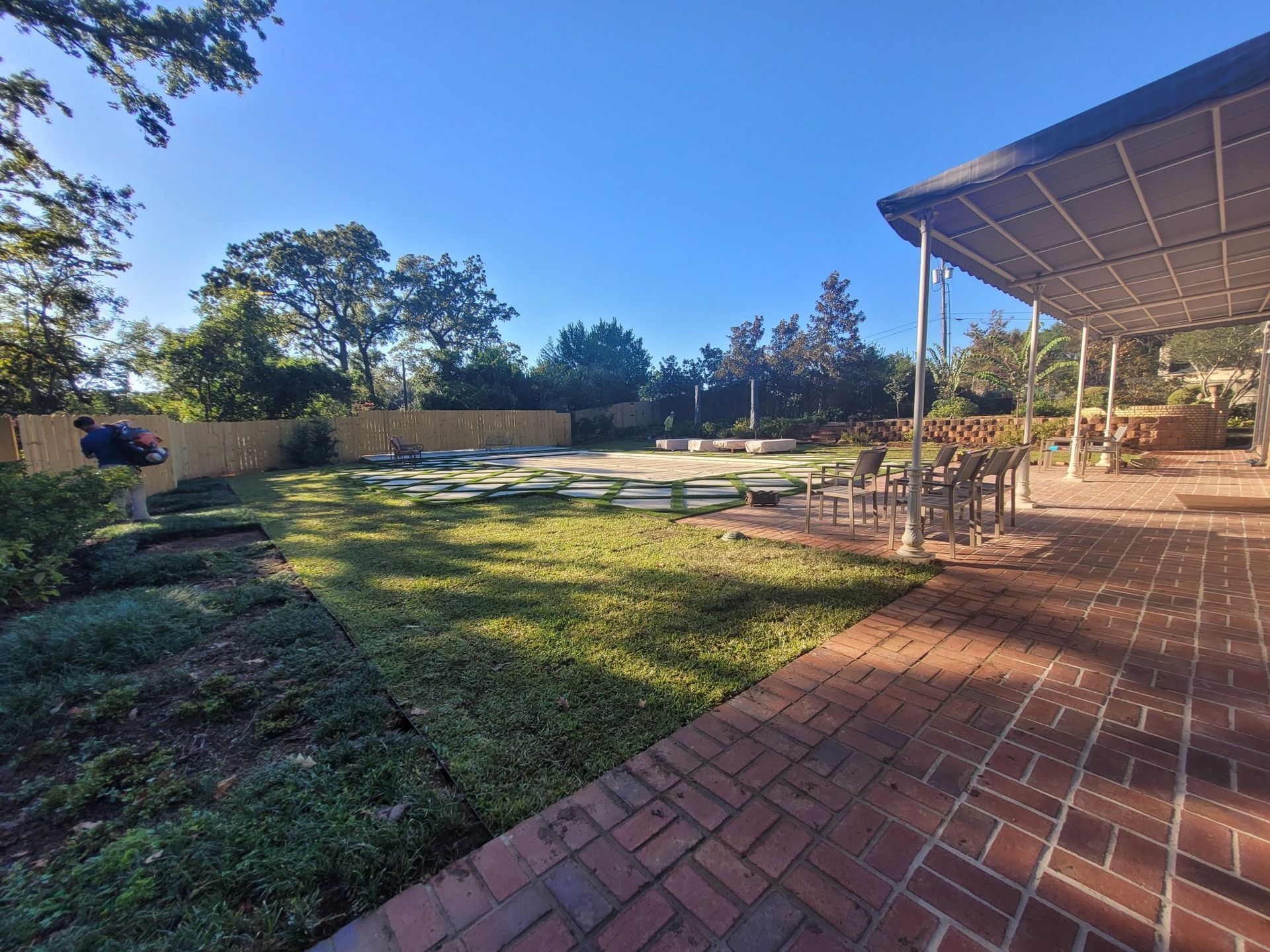 A person operates a leaf blower on a lawn next to a brick patio and a covered swimming pool on a sunny day.