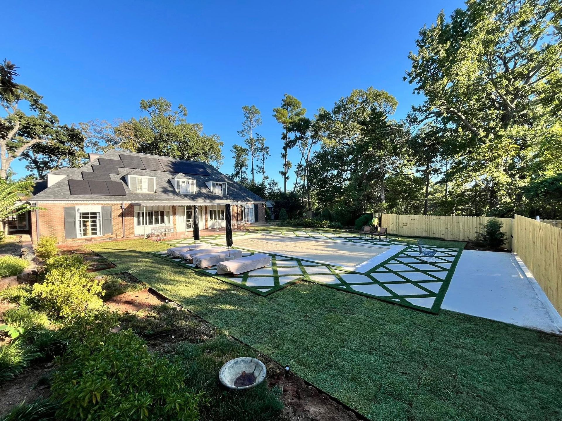 A brick house with a dark roof overlooks a backyard featuring a patio with a white lattice pattern and a pool.