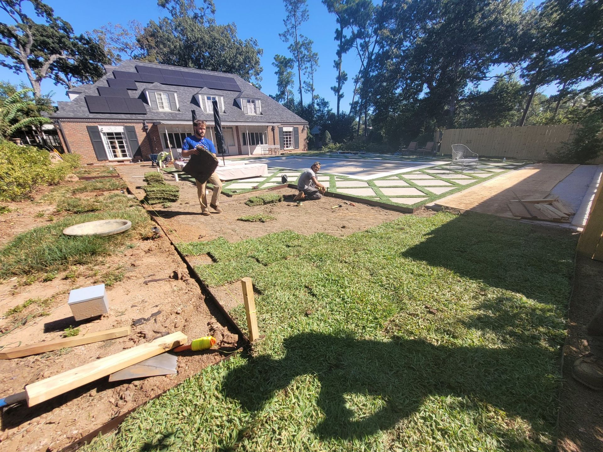 Two people work on landscaping a residential backyard featuring a modern brick home with solar panels and a patio.