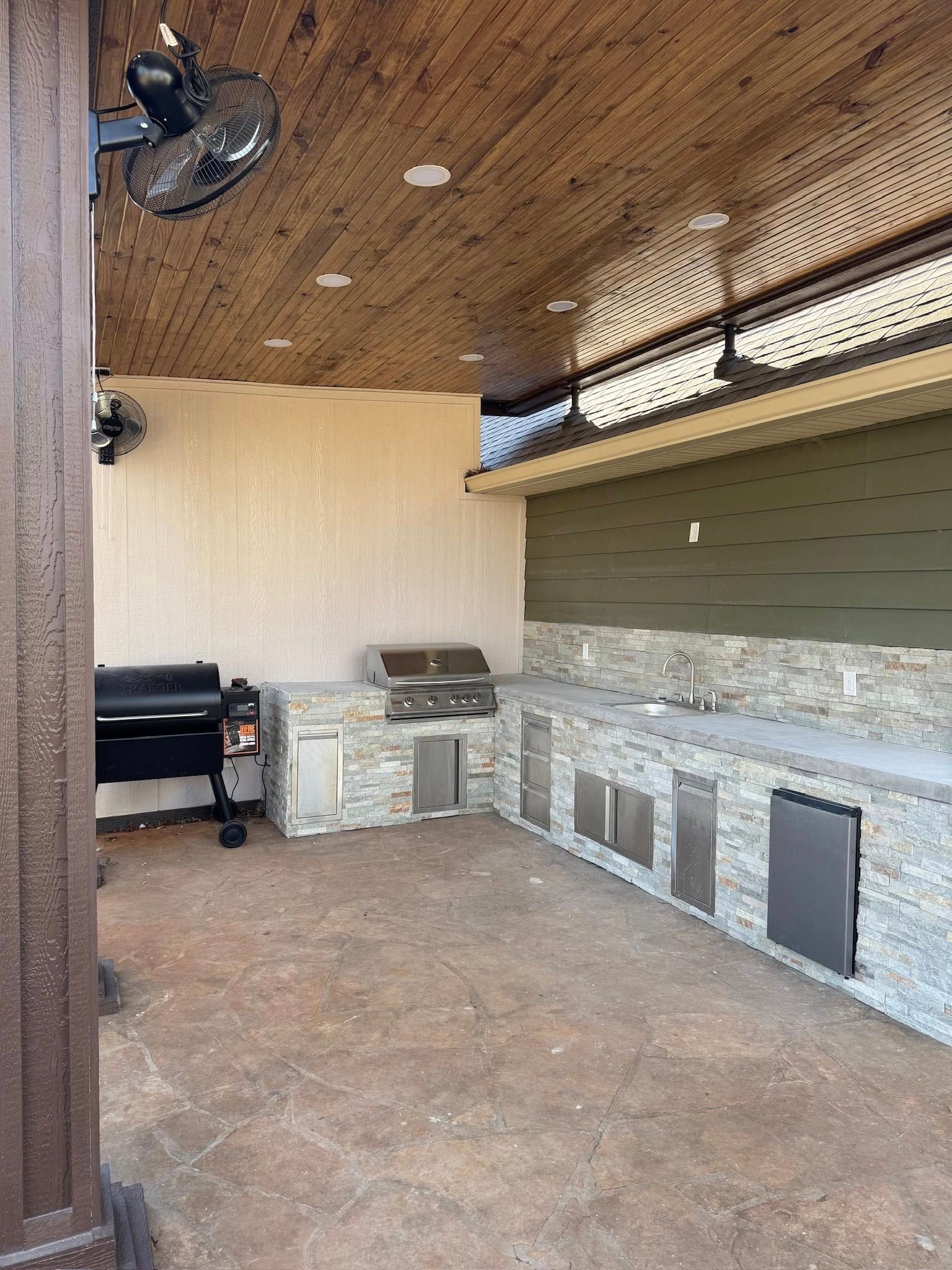 An outdoor kitchen with a stone counter, stainless steel grill and cabinets, dark wood ceiling, and a charcoal grill.