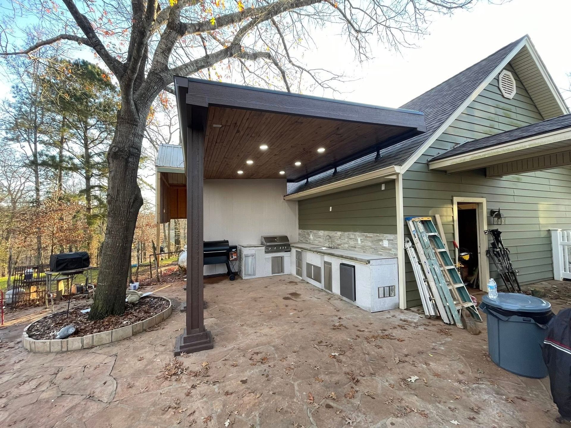 A partially constructed outdoor kitchen with a brown pergola roof and recessed lighting next to a green-sided house.