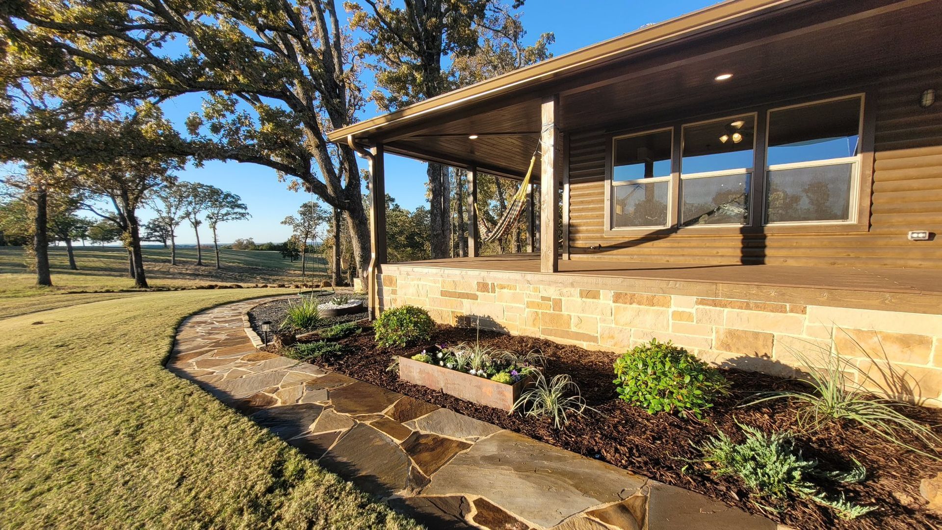 A wooden house with a stone porch and flagstone path surrounded by landscaping and trees under a clear blue sky.