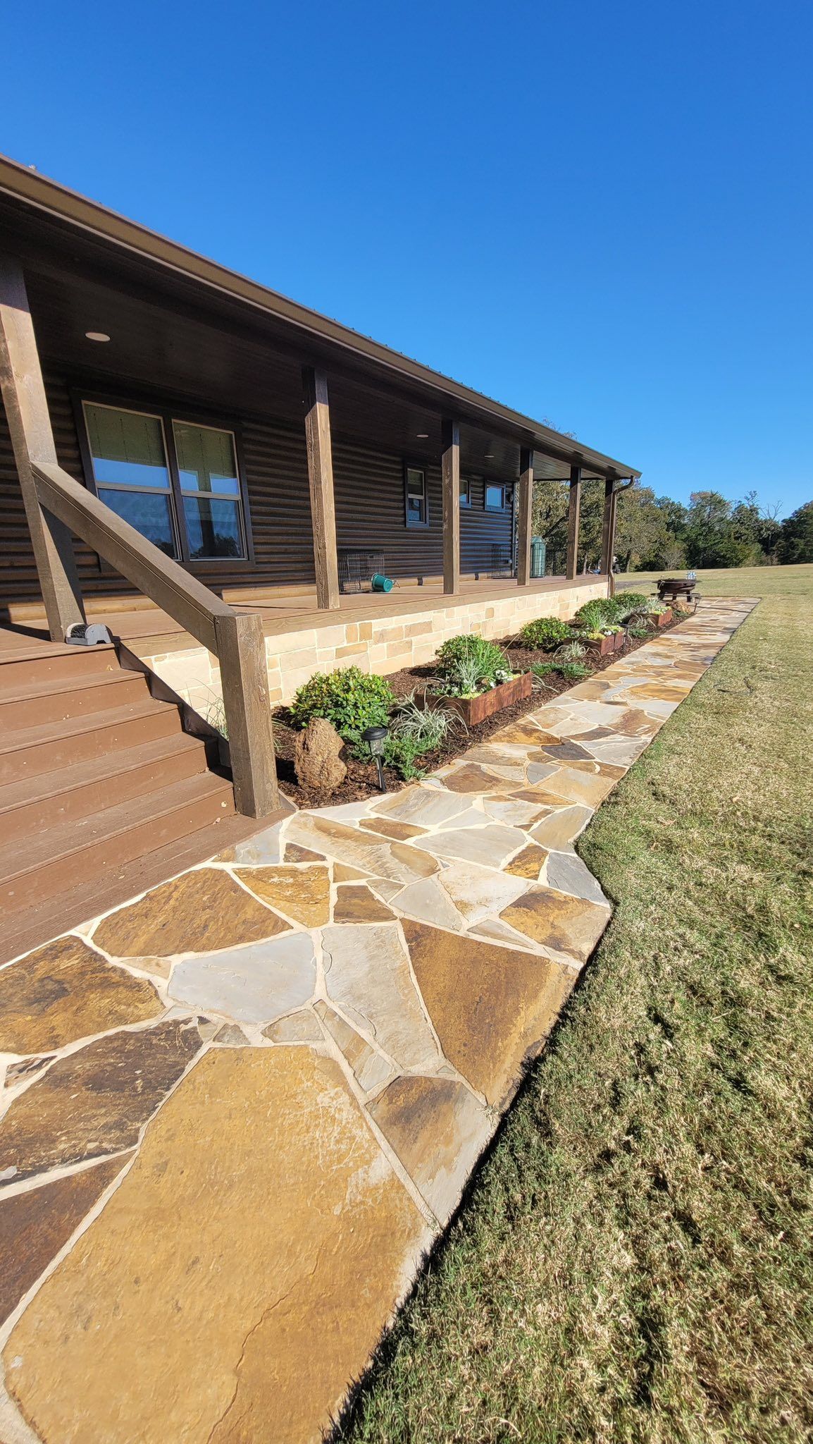 A log cabin with a wooden deck and stone walkway bordered by small plants under a clear blue sky.
