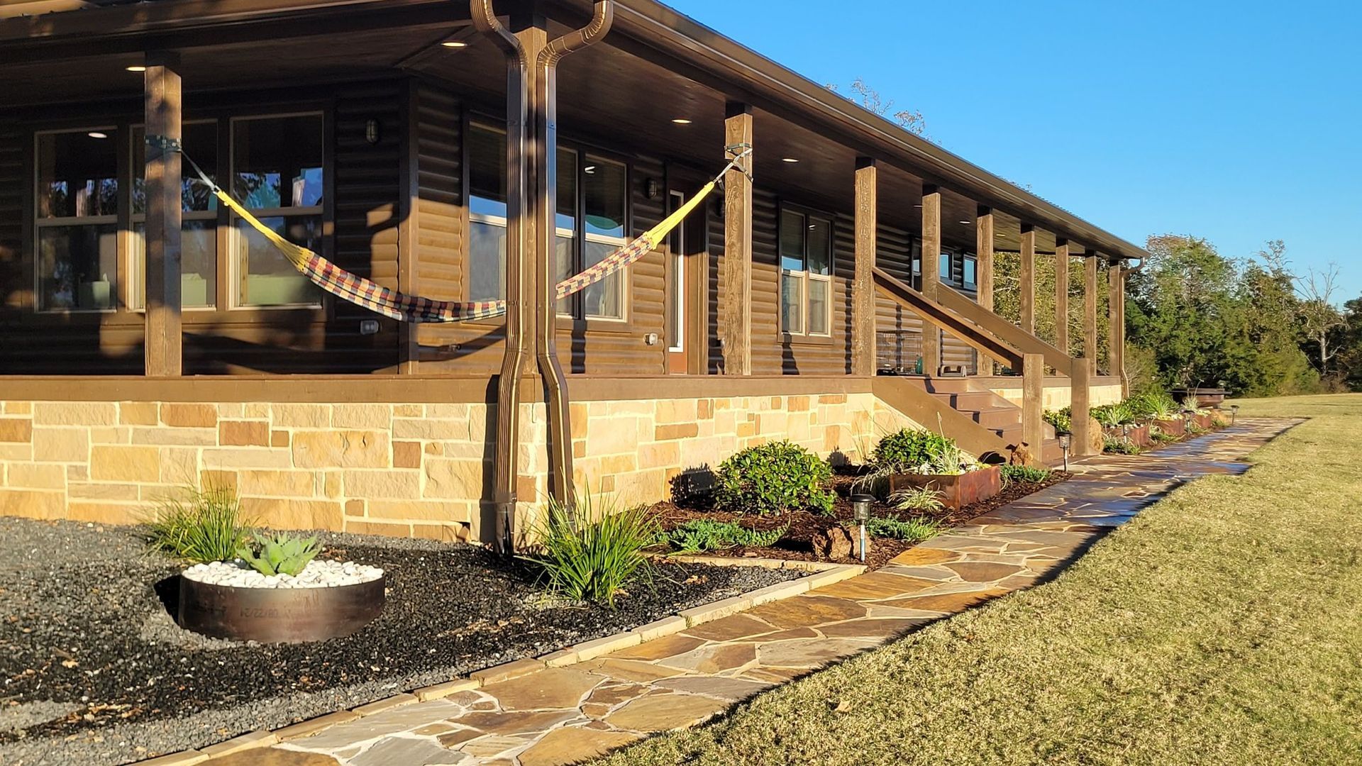 A dark wood log cabin with a stone foundation, a wide porch with a hammock, and a stone walkway beside a grassy yard.