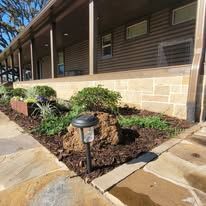 A solar path light stands in a mulch bed with a large rock and greenery in front of a house with stone siding and a porch.