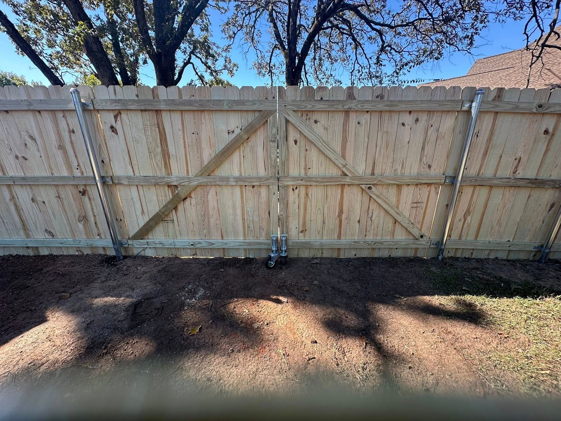 A double wooden fence gate with diagonal support beams, wheels at the bottom, and metal hinges against a sunny sky.