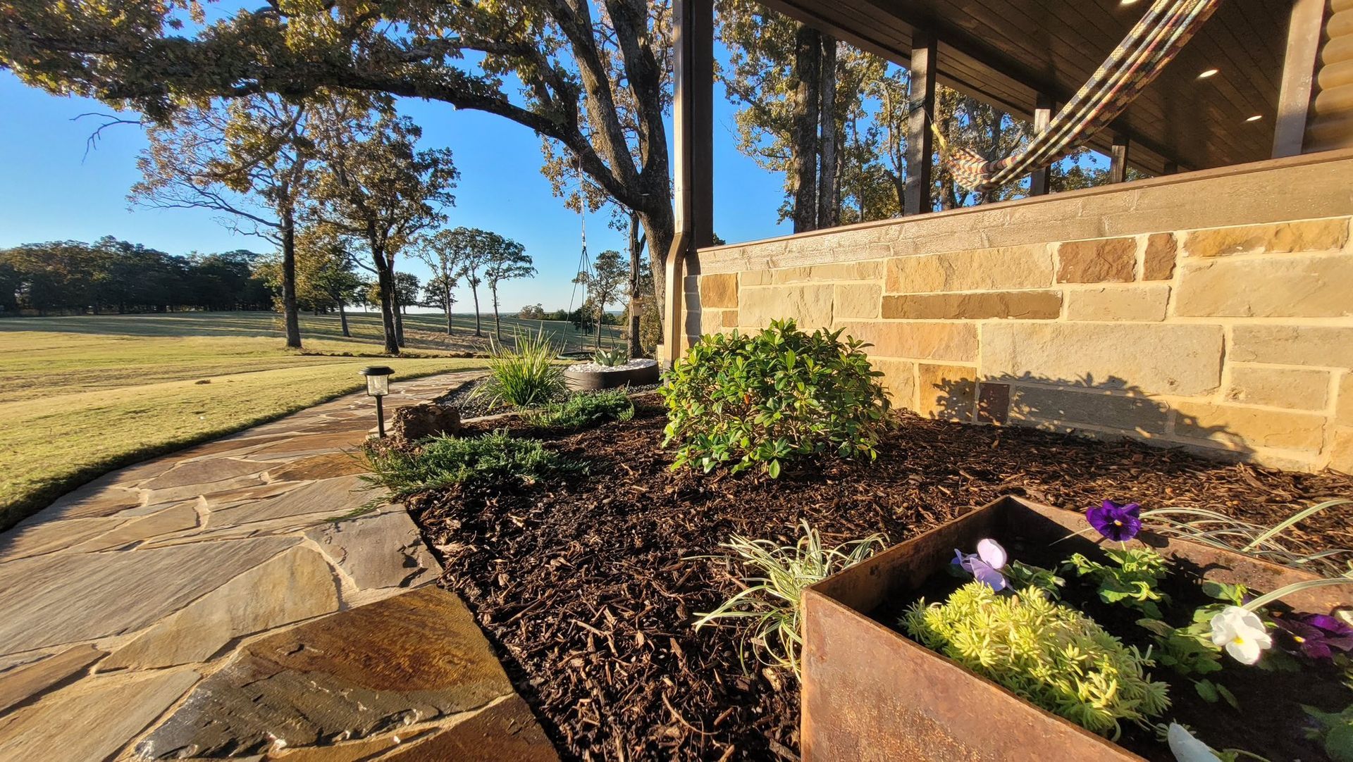A stone walkway leads past a garden bed with mulch and small shrubs toward a house with a stone porch on a sunny day.