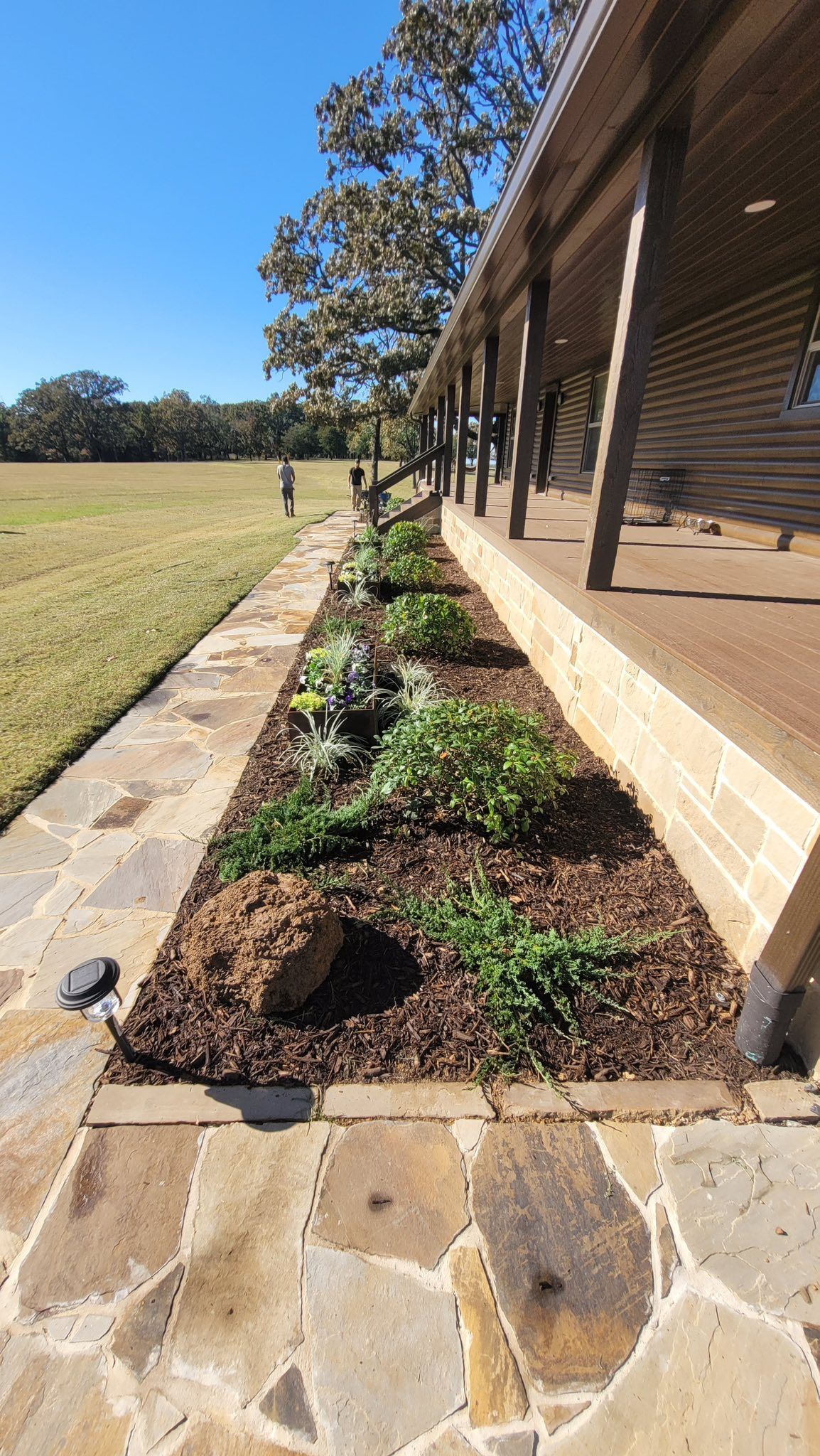 A stone pathway beside a building with a porch, featuring a mulched garden bed with small green plants and a large rock.