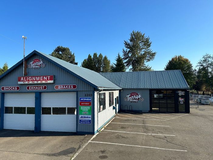 Gray auto repair shop with garage doors, red sign, and empty parking lot under a blue sky