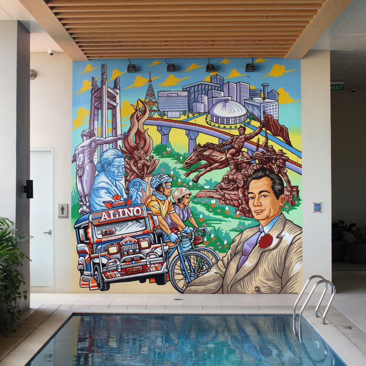 Colorful mural above an indoor pool, showing city landmarks, a bridge, and cyclists under a wooden ceiling