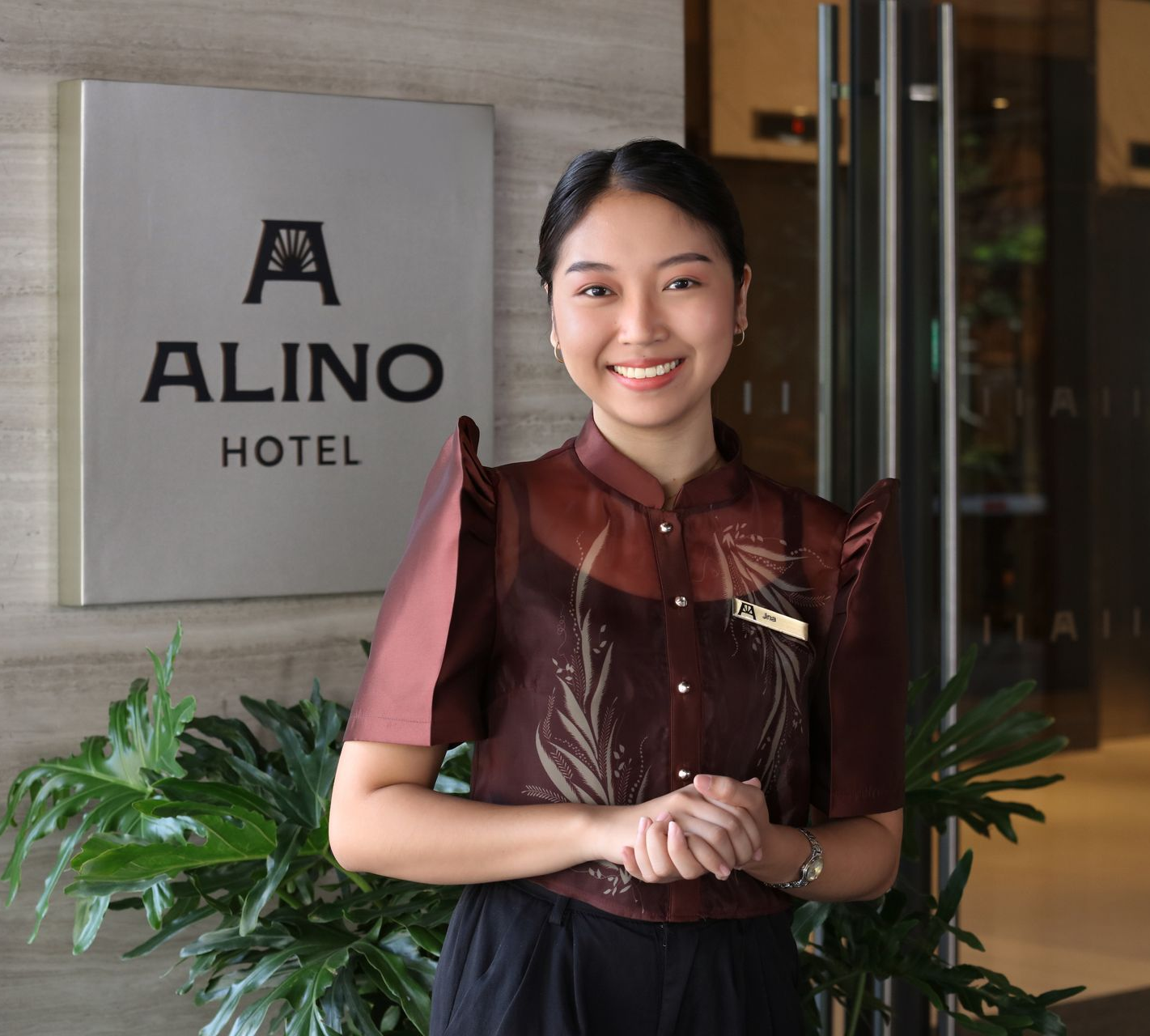 Hotel staff smiling beside an ALINO HOTEL sign, with plants in the lobby.