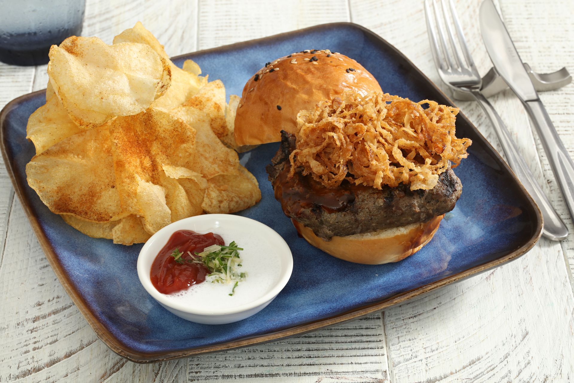 A blue rectangular plate holds a burger topped with fried onions, a side of potato chips, and a small dish of dip.