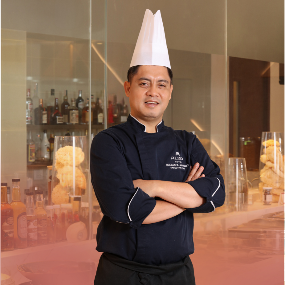 Chef in black uniform and white hat stands with arms crossed in a restaurant kitchen.